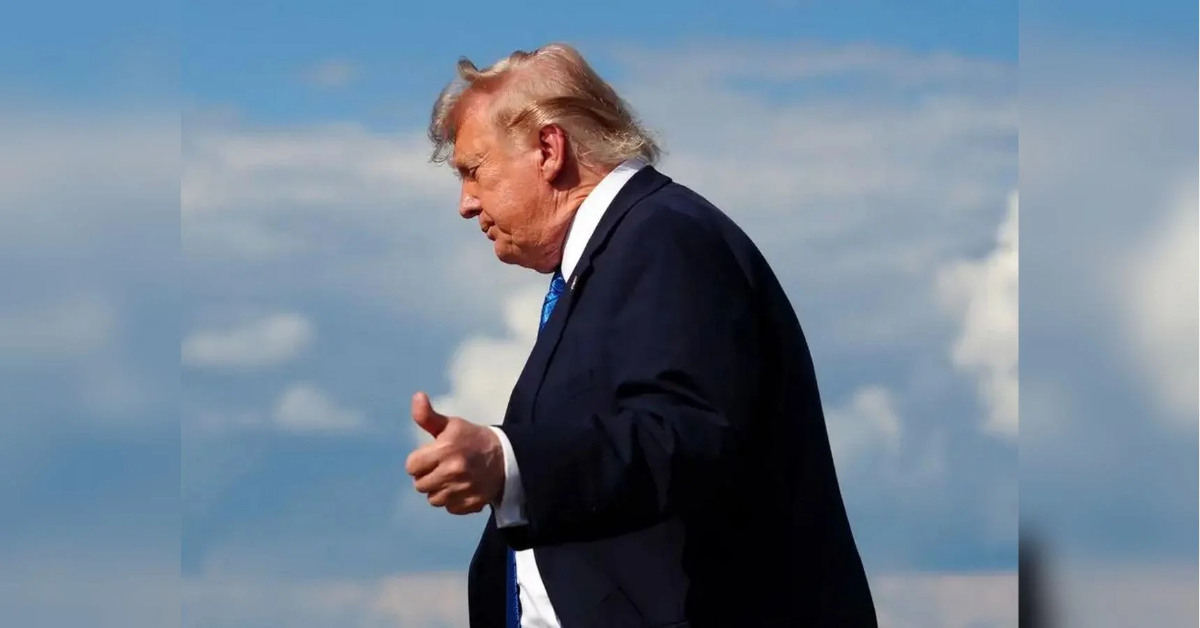 Дональд Трамп у темному костюмі показує великий палець вгору на тлі блакитного неба з хмарами